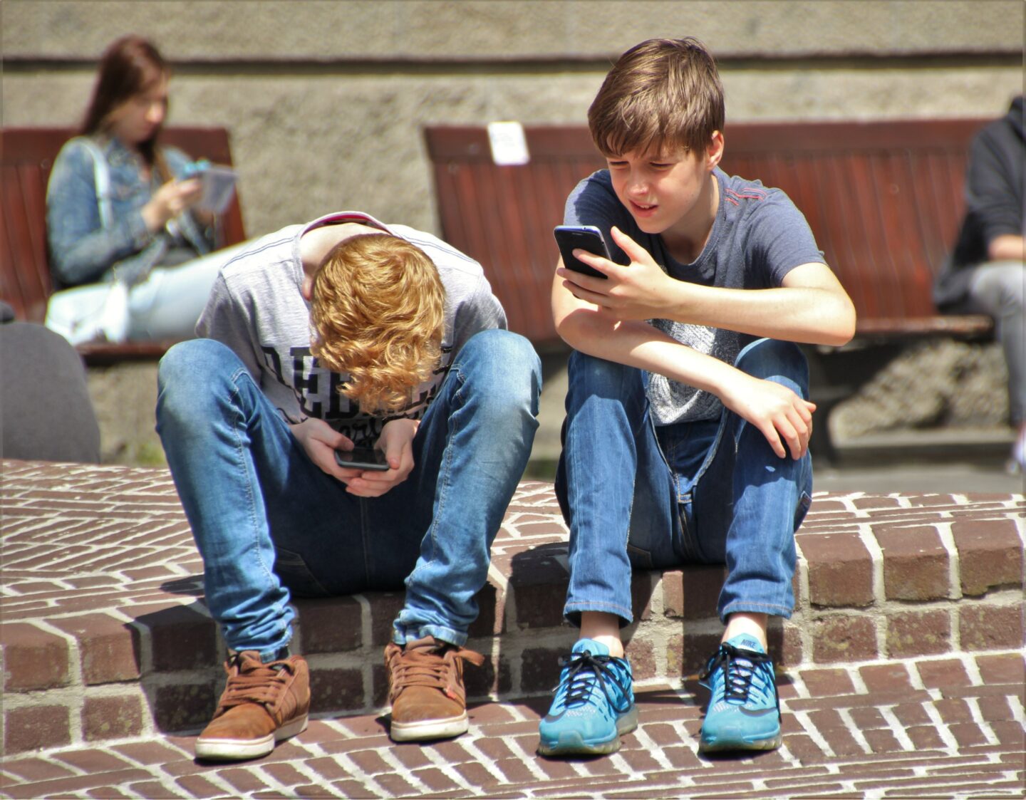 Two children sitting next to one another outside are fixated on their phones
